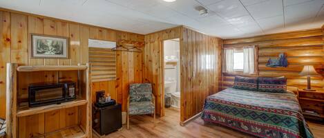 A wooden cabin room with a bed, a chair, a shelf with a microwave, and a picture on the wall.