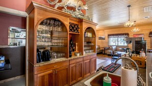 A wooden bar area with shelves of glasses and bottles, a mounted animal head, and a kitchen area with a stove and seating. - Sportsmans Lodge (Ennis)