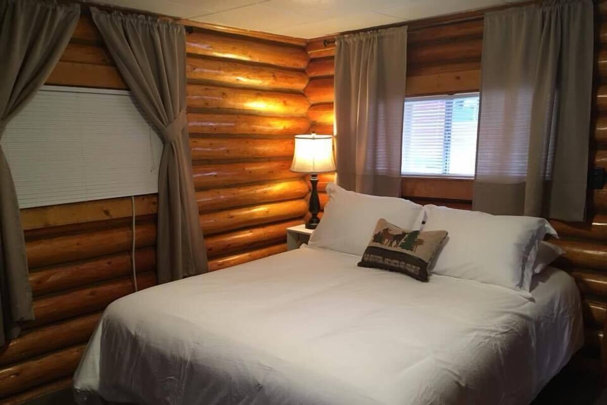 A log cabin-style room with a large bed, curtains, a window with blinds, and a lamp on a nightstand.