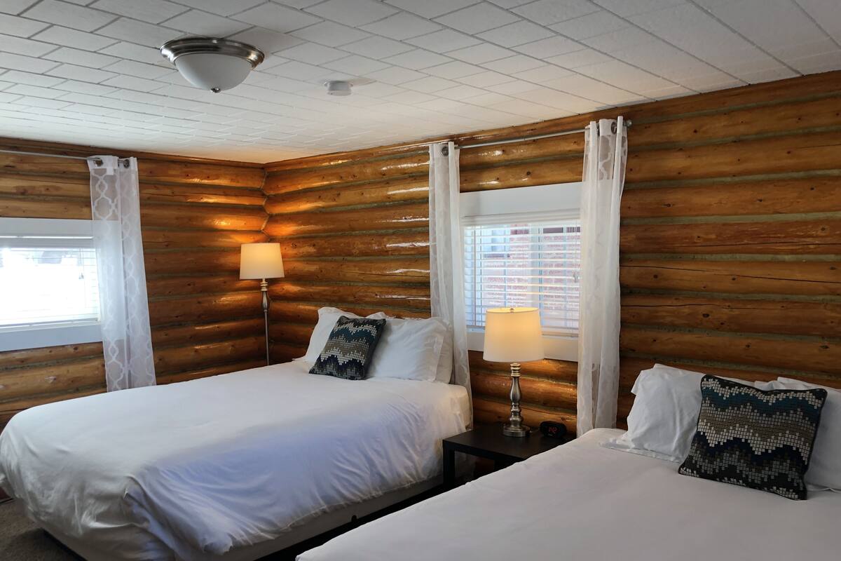 A room with two beds, wooden walls, a ceiling light, and a window with blinds.