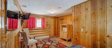 A log cabin room with a bed, a chair, a small table, a mirror, and a window with red curtains.