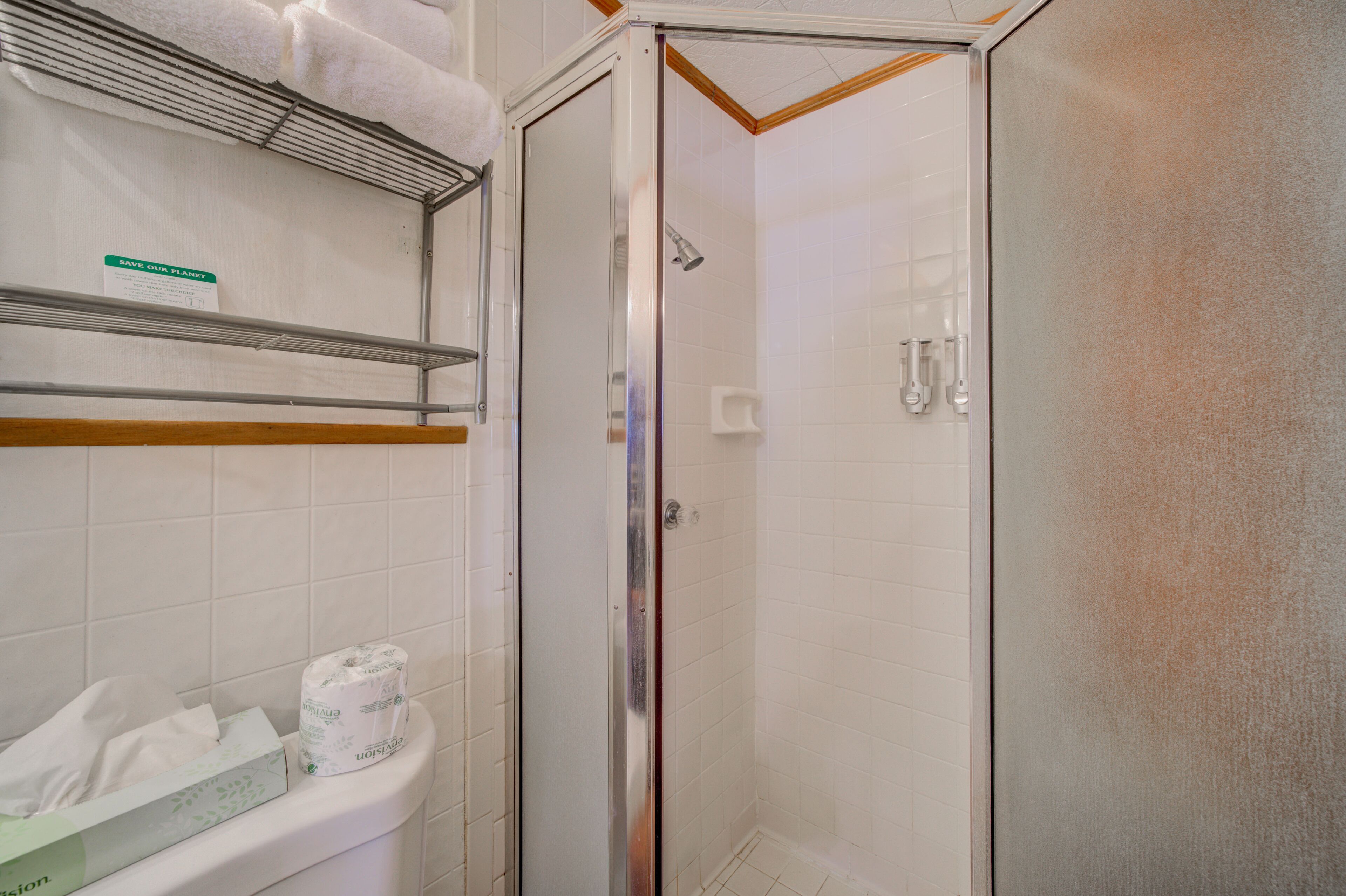 A shower area with a glass door, a toilet with a paper towel dispenser, and a shelf with towels.