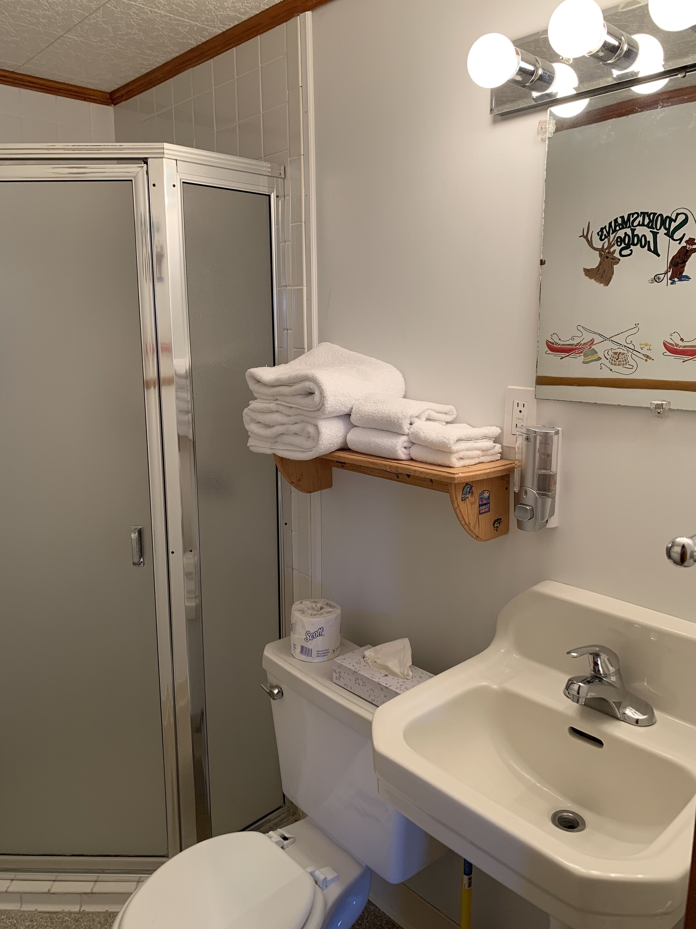 Un baño con inodoro, lavabo y zona de ducha. Hay toallas apiladas en un estante sobre el inodoro.