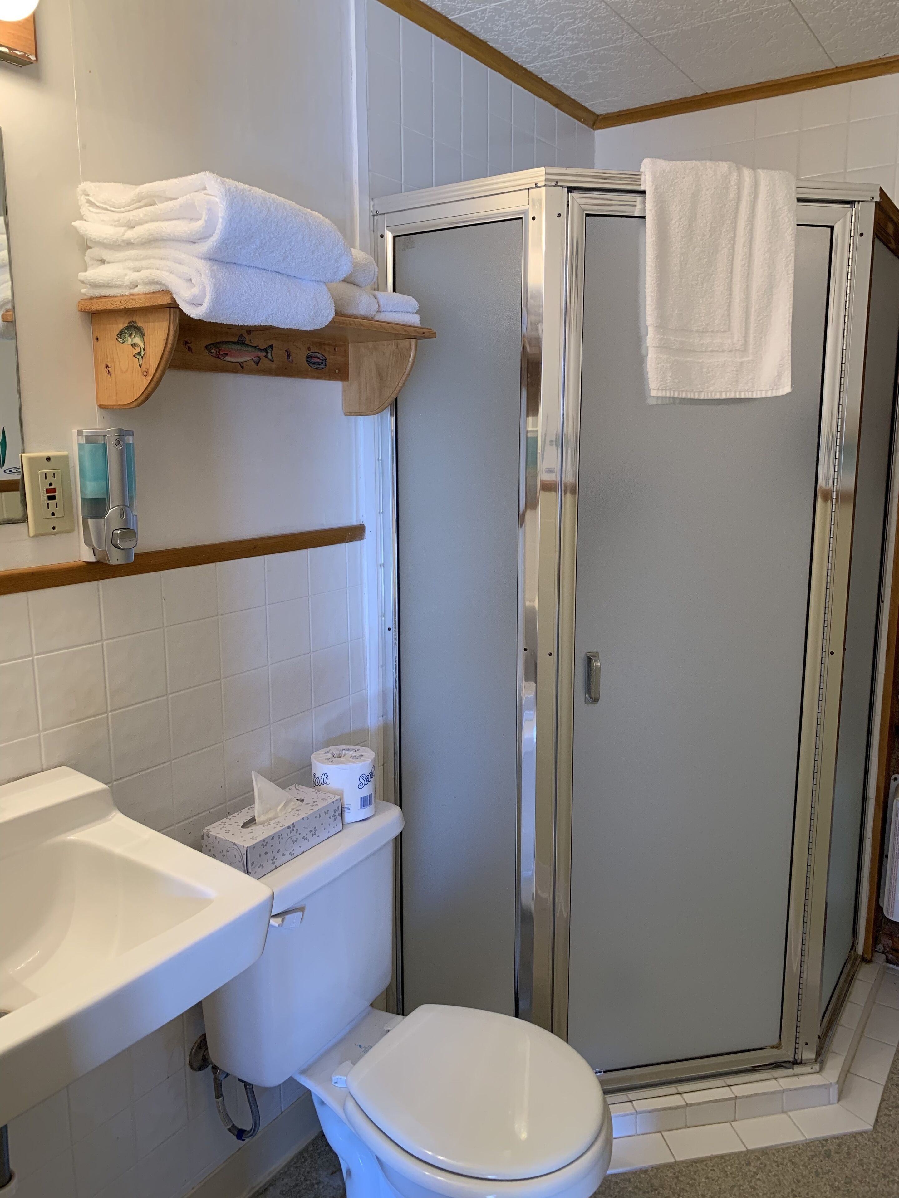 A bathroom with a white sink, toilet, and a glass-enclosed shower. There are towels stacked on a wooden shelf and a soap dispenser on the wall.