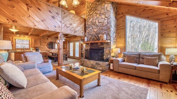 A cozy cabin interior with a stone fireplace, wooden walls, and a large window overlooking trees.