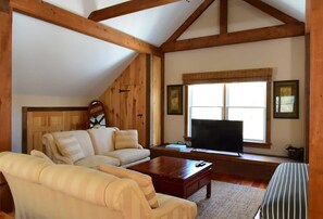 Un salon chaleureux avec un plafond haut, des poutres en bois, une grande fenêtre avec des stores, un téléviseur à écran plat et un canapé.