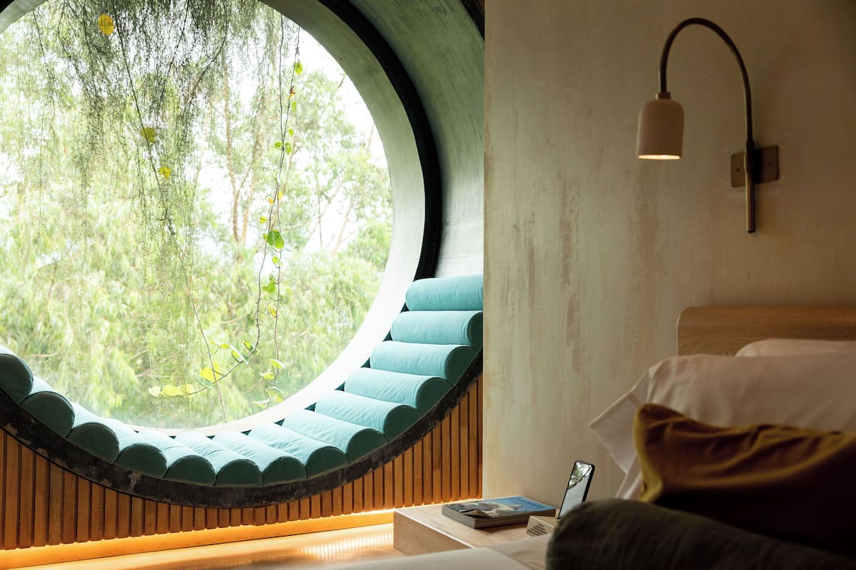 A room with a circular window, a bed with a wooden headboard, a nightstand with a lamp, and a view of greenery outside.
