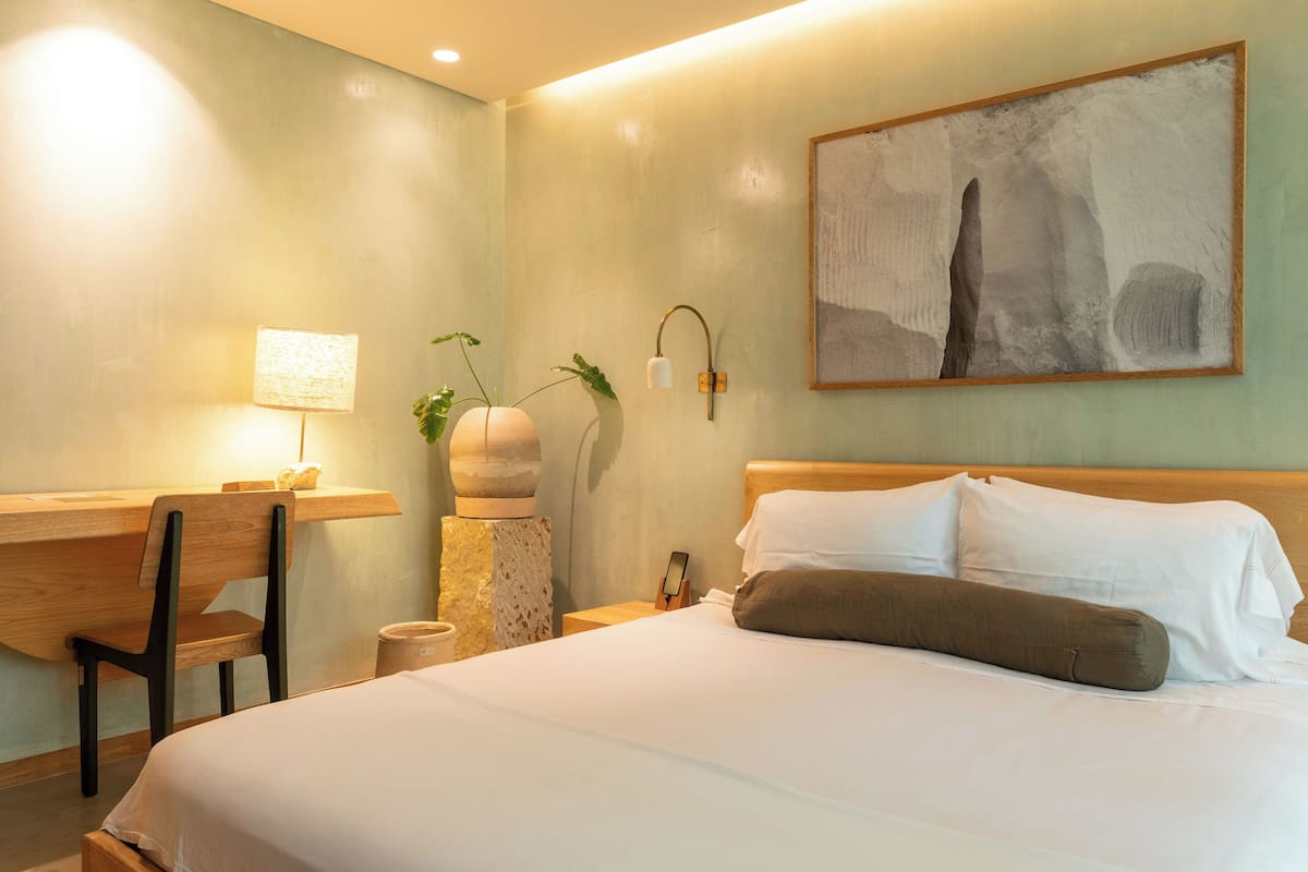 A hotel room with a bed, a desk, a chair, a lamp, a wall-mounted phone, and a painting.