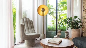 A modern living room with a stone bench, a round wooden table with a book, a floor lamp, and a view of greenery through large windows. - 23Hotel Medellin (Medellín)