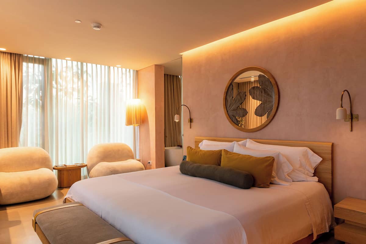 A modern hotel room with a large bed, two armchairs, a round mirror, and a nightstand.