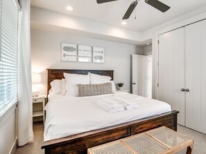 Quarto bem arrumado com uma cama de madeira, roupa de cama branca, mesa de cabeceira com abajur e um ventilador de teto.
