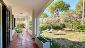 A covered patio with a tiled floor, a green chair, and a view of a garden with trees and sun loungers.