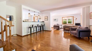 A spacious living area with a wooden staircase, a kitchen with bar stools, a grey sofa, and a television.