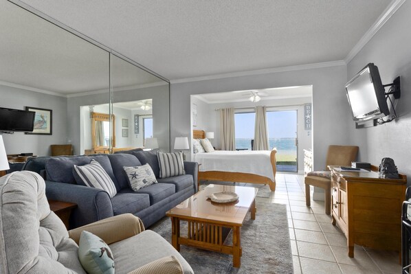A hotel room with a bed, a sofa, a coffee table, a TV, and a view of the sea. - Sunset Harbor Palms 1-102 | Sound Front | Patio, Gazebo, Pier, BBQ Grills (Navarre Beach)