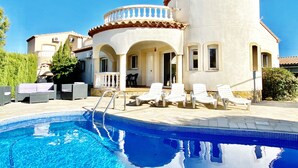 Un espace aménagé au bord d’une piscine à l’eau d’un bleu limpide, avec des chaises longues blanches et un bâtiment à la façade blanche et au toit recouvert de tuiles rouges.