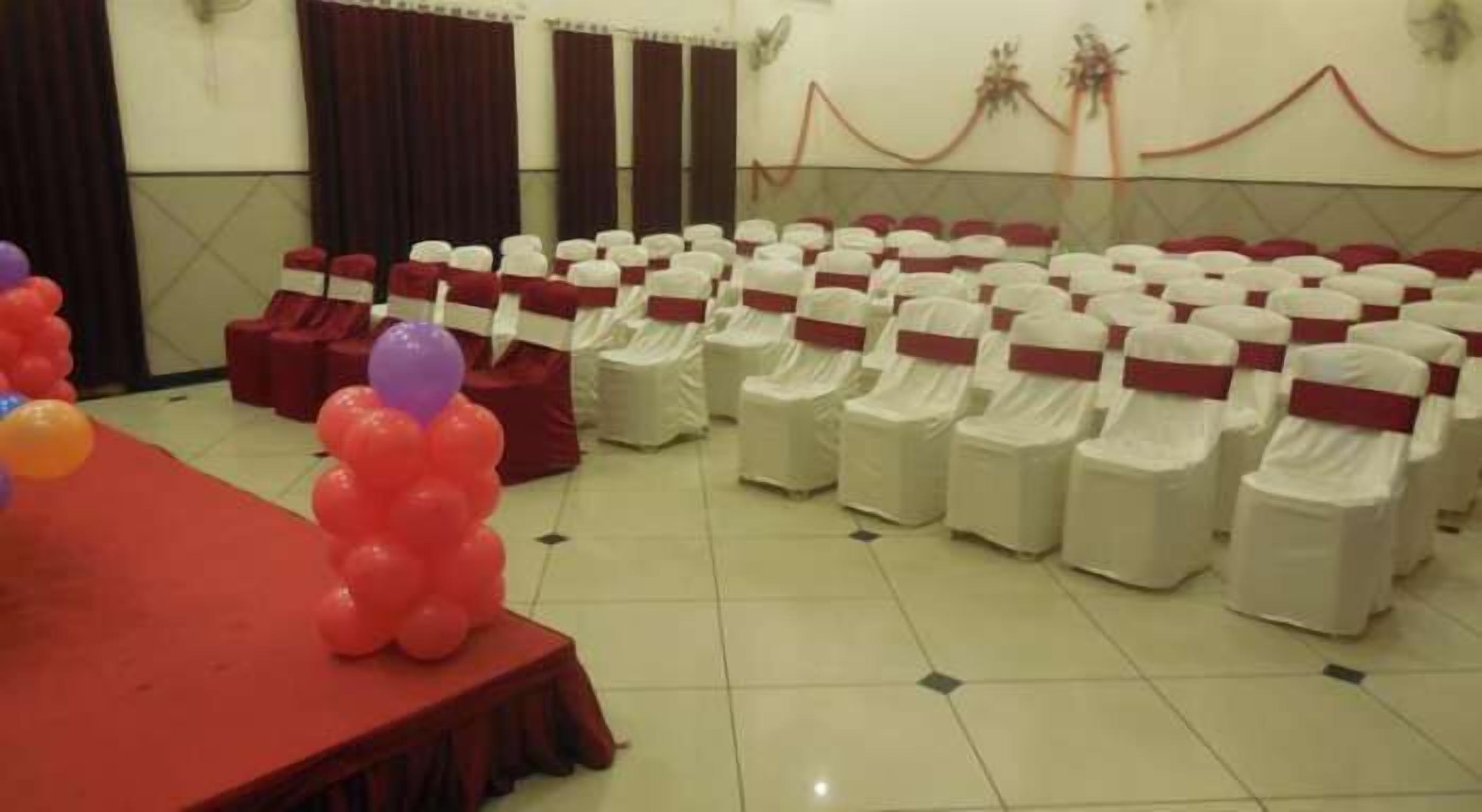 Une salle de conférence avec des rangées de chaises recouvertes de rubans blancs et rouges, une scène recouverte d’un tapis rouge et des décorations en ballons.