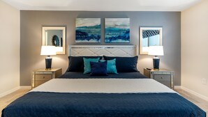 Un dormitorio con una cama grande, dos mesitas de noche con lámparas, un cabecero con espejo y dos cuadros abstractos en la pared.