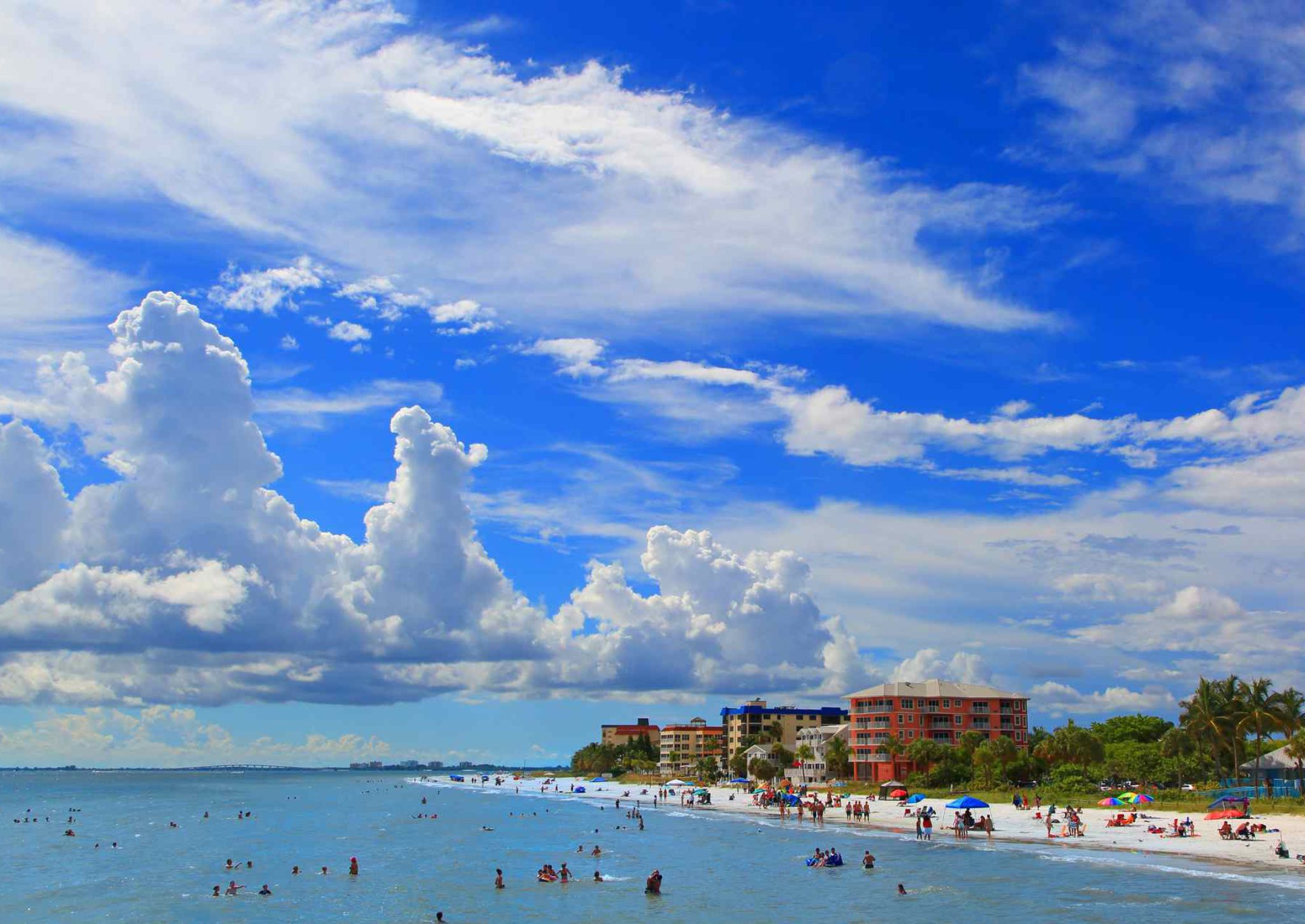 A beach with people swimming and sunbathing, a clear sky, and buildings in the background.