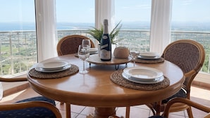 Una mesa redonda de madera con una botella de vino, dos copas y platos para dos personas, rodeada de sillas de mimbre, con vistas a un paisaje a través de ventanas amplias.