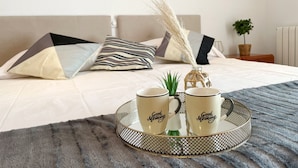 Una cama bien hecha con almohadas estampadas, una planta en la mesita de noche y una bandeja con dos tazas sobre una alfombra texturizada.