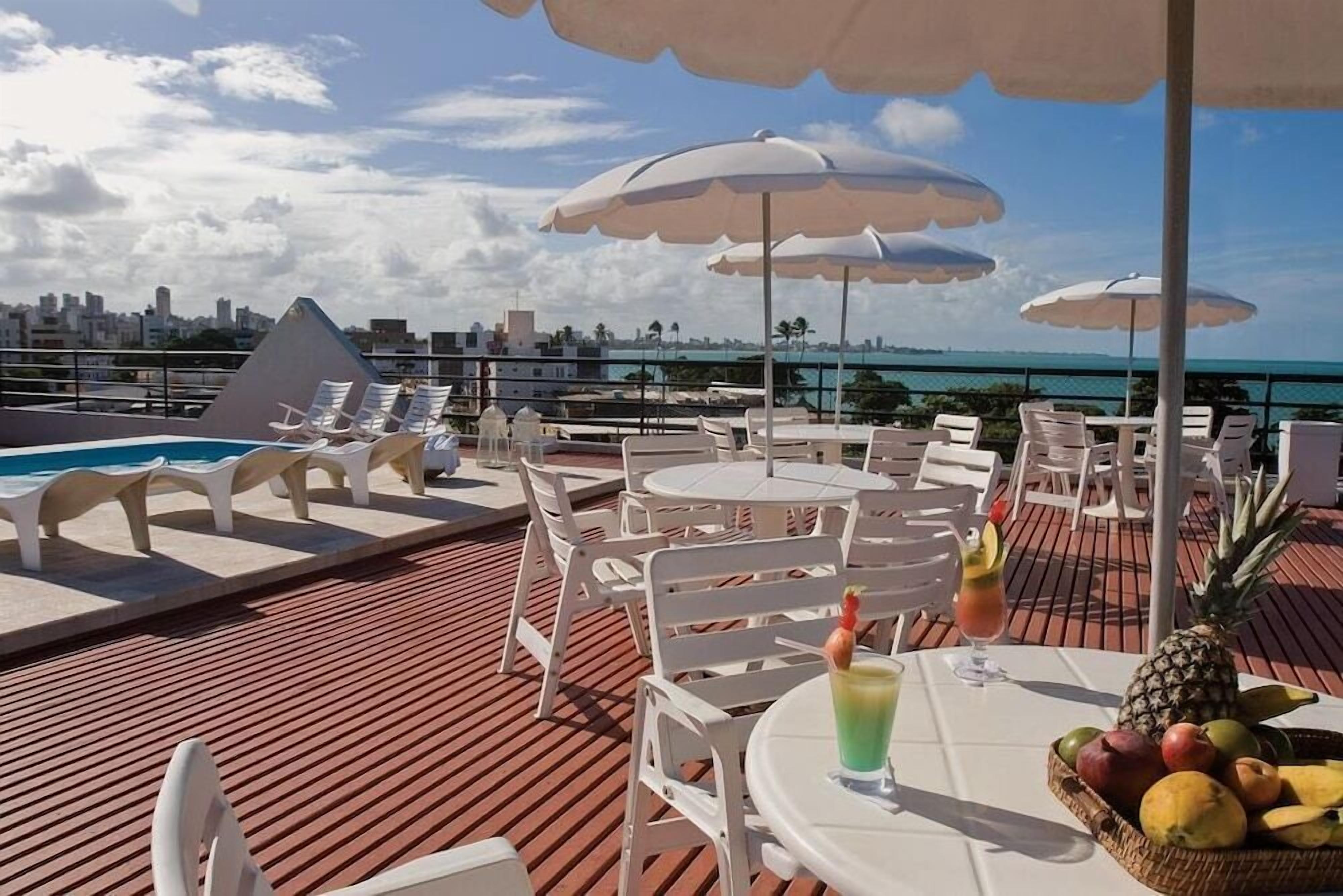 A rooftop bar with white chairs and tables, umbrellas, a pool, and a city view.