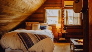 Un dormitorio acogedor en el ático, con techo inclinado de madera, una cama con manta a rayas, una guitarra y una mesita con lámpara.