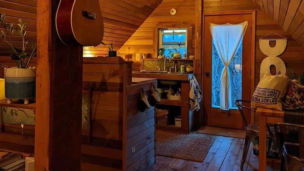 Interior acogedor de una cabaña de troncos, con una guitarra, paredes de madera, una zona de cocina y una ventana con cortinas.
