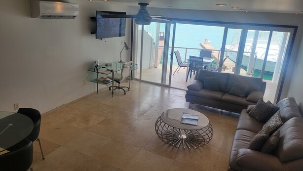 Ein Wohnzimmer mit Glastisch, einer Couch, einem Fernseher und einem Balkon mit Meerblick.