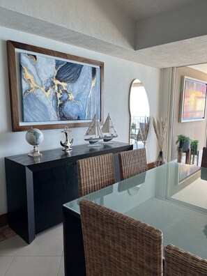 Un comedor moderno con una mesa de vidrio, sillas de mimbre, un aparador oscuro con objetos decorativos y un cuadro abstracto grande en la pared.