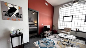 A modern living room with a red accent wall, a large window with blinds, a round coffee table, a sofa, and a flat-screen TV. - Stay Roanoke | Luxe Loft | Arts & Ale (Roanoke)