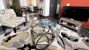 A modern living room with a sofa, a coffee table, a TV stand, and a dining area in the background. - Stay Roanoke | Luxe Loft | Arts & Ale (Roanoke)