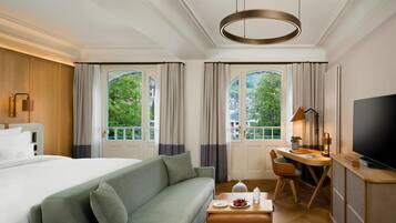 Habitación de hotel con cama, sofá verde, escritorio con silla, televisor y ventanas grandes con cortinas.