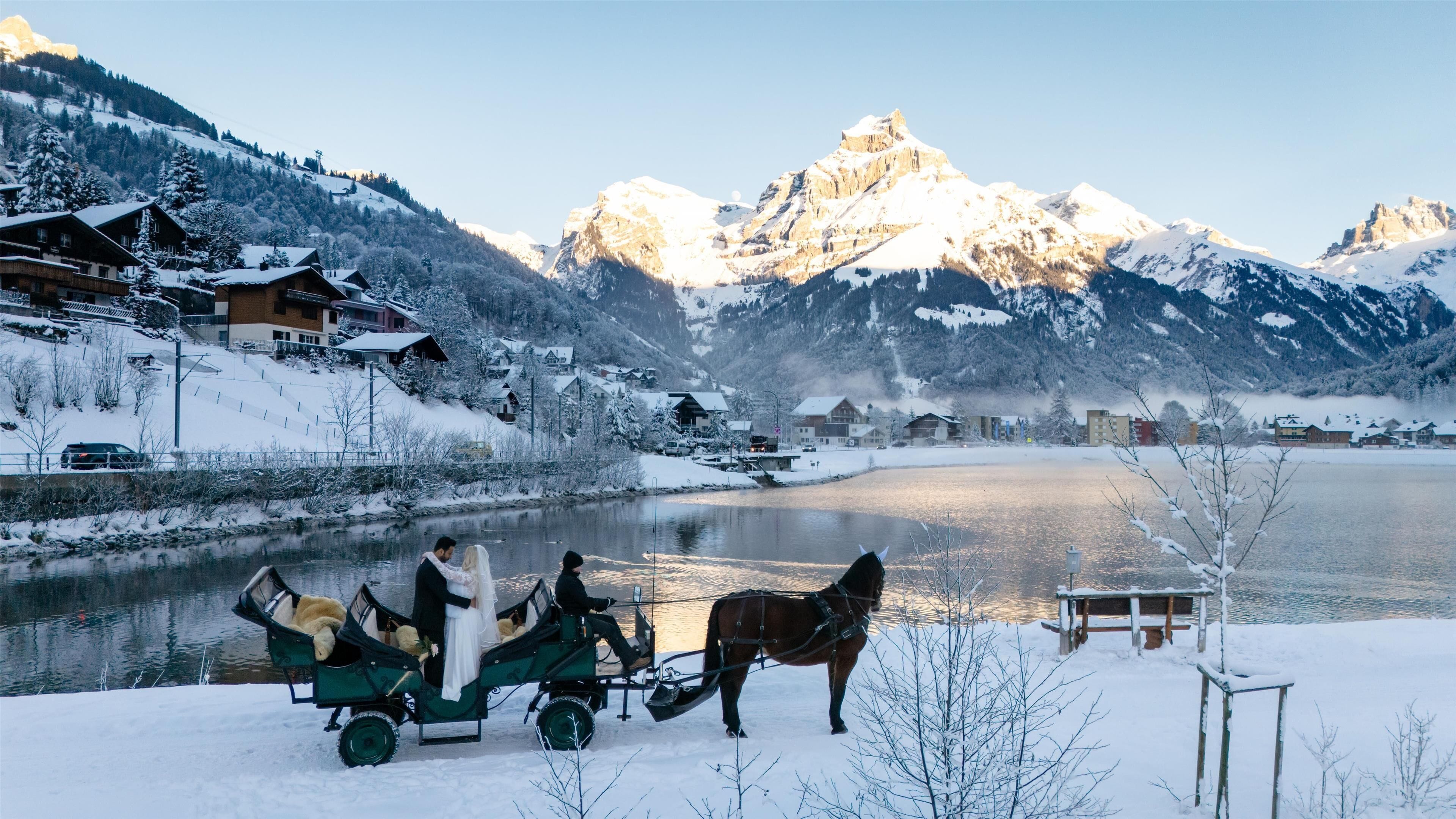 Um lago coberto de neve com uma carruagem puxada por cavalos e um casal com trajes de casamento.