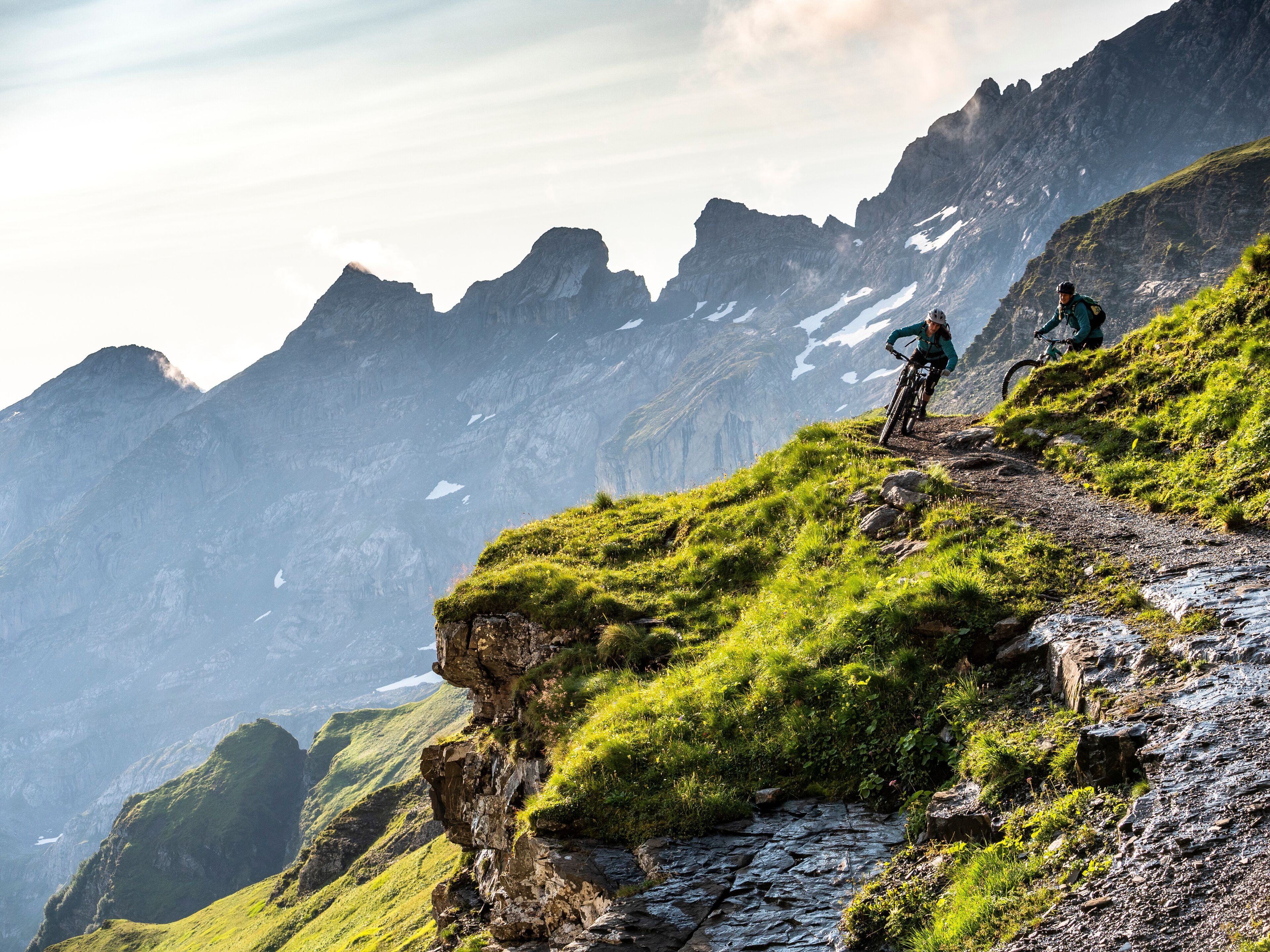 A mountain biking trail with rocky terrain and grassy areas, surrounded by steep cliffs and a valley.