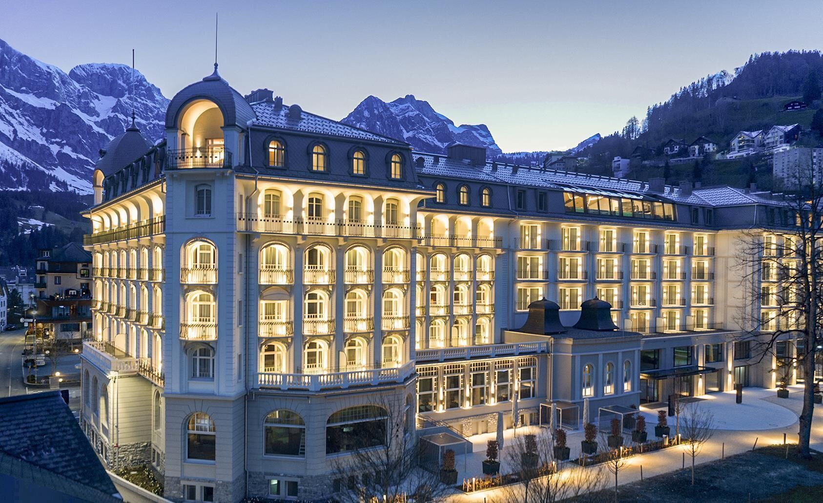 Un grand hôtel à plusieurs étages, de style architectural européen classique, avec des fenêtres en arc et des balcons, le tout situé dans un cadre montagneux.