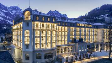 Un grand hôtel à plusieurs étages, de style architectural européen classique, avec des fenêtres en arc et des balcons, le tout situé dans un cadre montagneux.