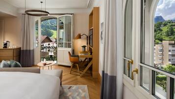 A hotel room with a bed, a desk, a chair, a lamp, and a view of a mountainous landscape.