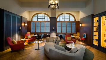 A spacious lounge area with a large sofa, armchairs, and a coffee table. There are two large arched windows providing natural light, and a chandelier hanging from the ceiling.