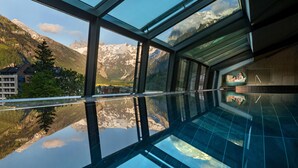 Una piscina infinita con paredes de vidrio, vista a la montaña, una terraza de madera y un área de descanso con una silla.