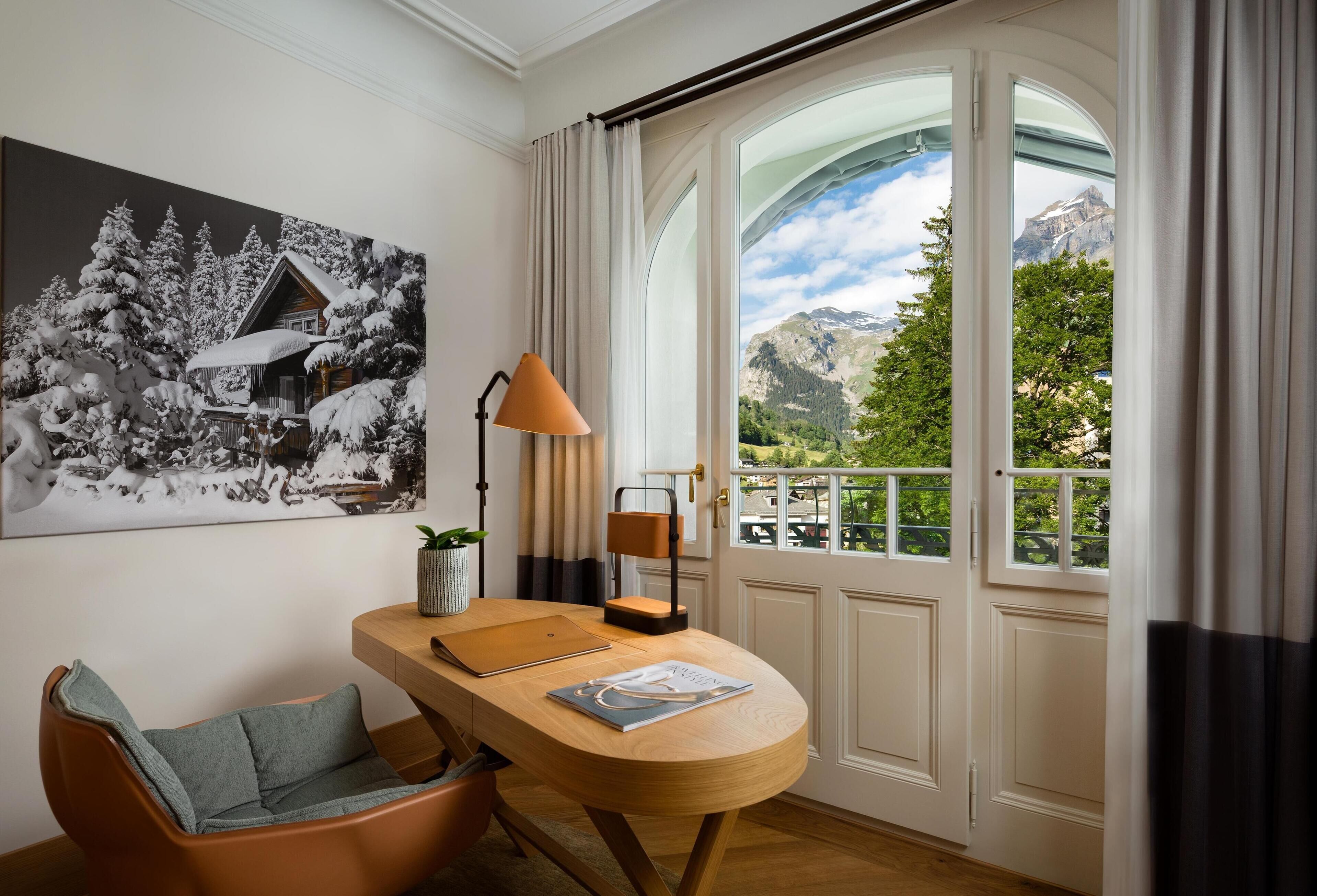 Una habitación con un escritorio de madera, una silla, una lámpara, un cuadro enmarcado y un amplio ventanal con vistas a la montaña.