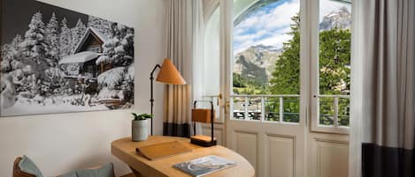 A room with a wooden desk, a chair, a lamp, a framed picture, and a large window with a mountain view.