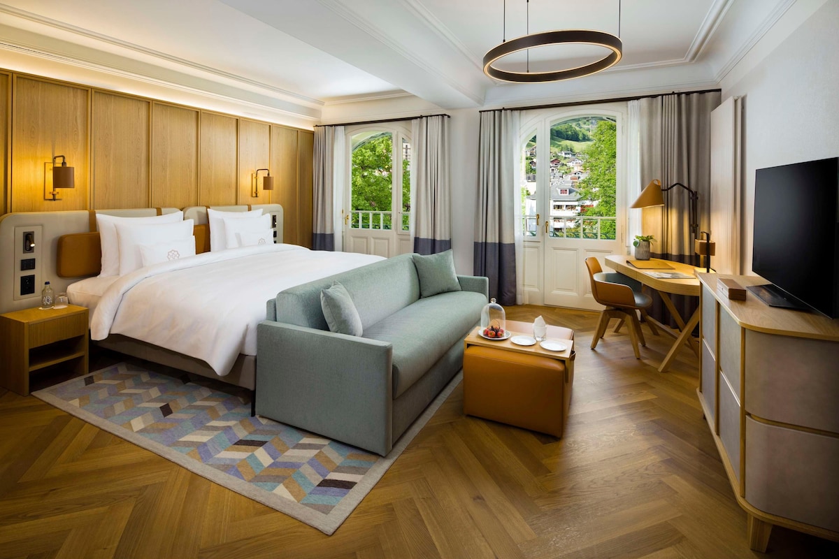 A hotel room with a large bed, a sofa, a desk, a TV, and a view of greenery.