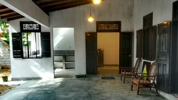Un espacio amplio y cubierto con suelo de hormigón, techo de madera y una lámpara colgante. Varias sillas de madera y una puerta con paneles decorativos.
