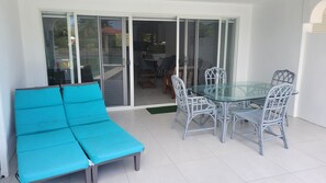Eine Terrasse mit einem Glastisch, Korbstühlen, einem blauen, gepolsterten Sofa und einer Glastür, die in einen Innenbereich mit einem Esstisch und Stühlen führt.