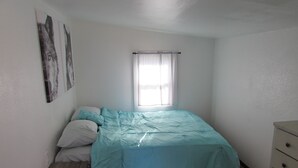 A small, neatly arranged bedroom with a bed, a nightstand, a dresser, and a window with sheer curtains. - Teakettle Kottage (Columbia Falls)