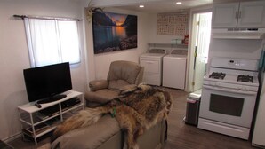 A living room with a television, a sofa, a dog, a painting, a kitchen with appliances, and a window with curtains. - Teakettle Kottage (Columbia Falls)