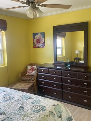Un dormitorio con cama, cómoda, silla, ventilador de techo y ventana.