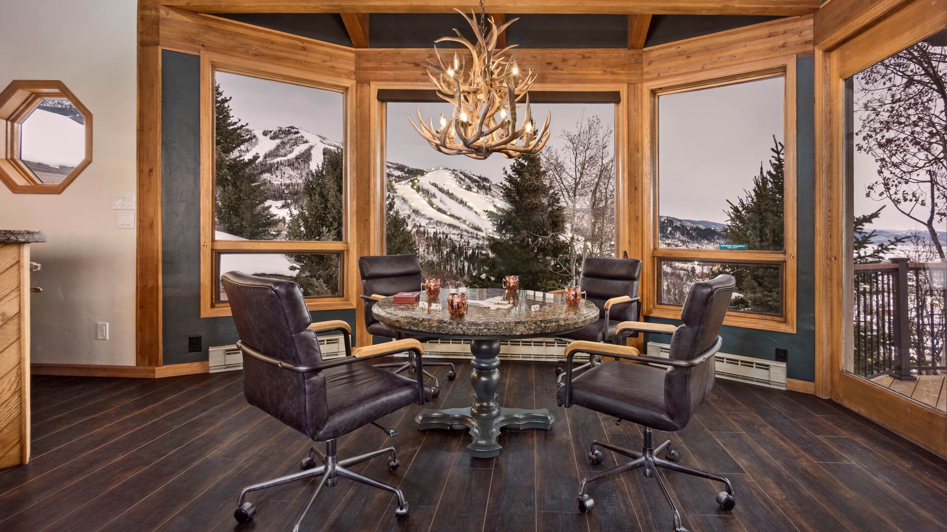Una habitación acogedora con una mesa redonda, cuatro sillas y un candelabro. Amplias ventanas ofrecen vistas de montañas nevadas.