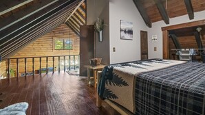 Une pièce chaleureuse avec un plafond en bois, un lit recouvert d’une courtepointe à motifs, une petite table sur laquelle est posé un vase, et une fenêtre donnant sur un paysage verdoyant.