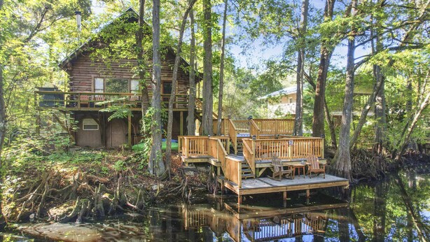 Une cabane en bois avec une jetée qui s’avance dans une zone marécageuse, entourée de grands arbres et dont le reflet se mire dans l’eau.